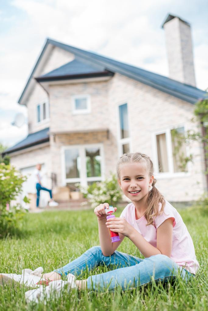 очаровательный маленький ребенок с мыльными пузырьками, сидящий на газоне, в то время как ее мать стоит позади перед домом
  - Фото, изображение