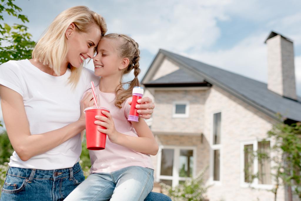 glückliche Frau sitzt Stirn an Stirn mit Tochter und hält Blasengebläse vor dem neuen Haus - Foto, Bild