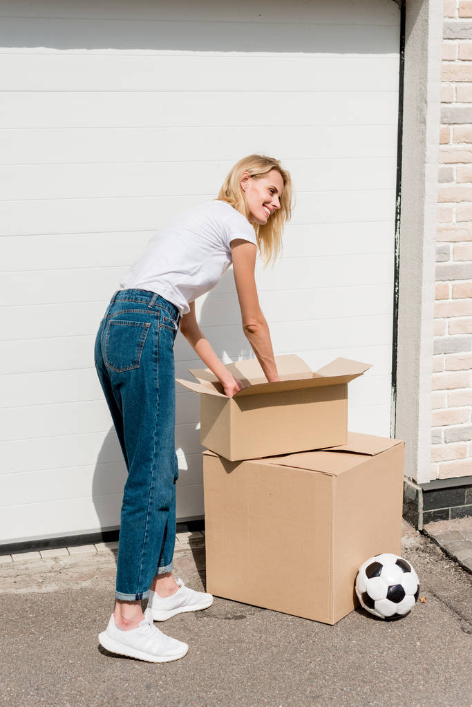  mujer joven desempacando cajas de cartón cerca de la pelota de fútbol en frente del garaje de la nueva casa
  - Foto, imagen