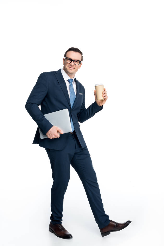full length view of handsome businessman holding laptop and paper cup and smiling at camera isolated on white  - Photo, Image