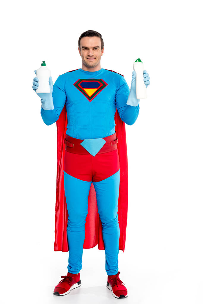 pohledný muž v kostýmu superhrdiny uchycení lahví s mycím prostředkem a usmívá se na kameru izolované na bílém  - Fotografie, Obrázek