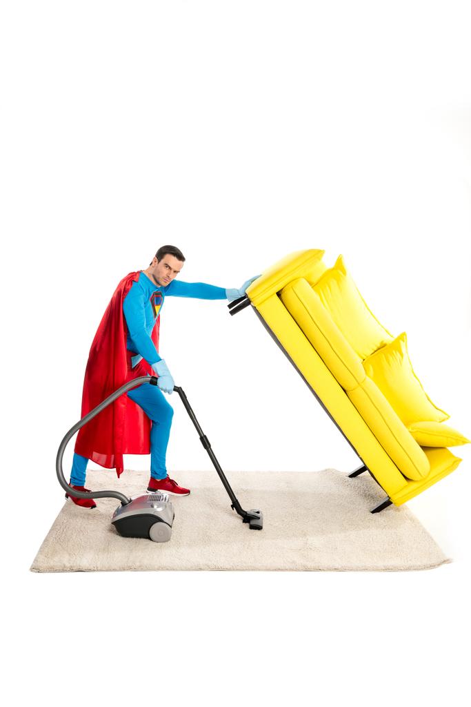 male superhero holding couch while cleaning carpet with vacuum cleaner and looking at camera isolated on white - Photo, Image