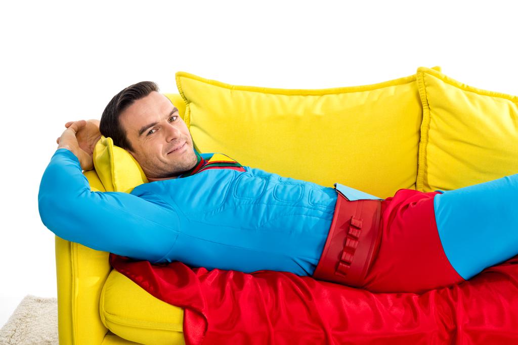 szép ember a szuperhős jelmezben pihent a kanapén, és mosolyogva elszigetelt fehér kamera - Fotó, kép