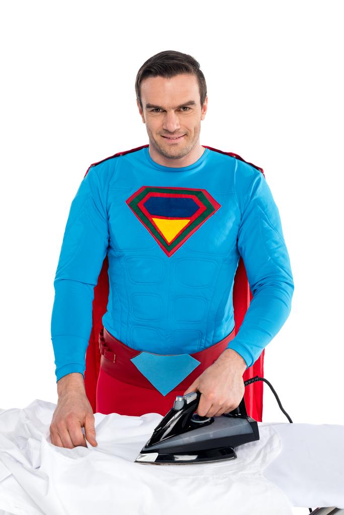 pohledný muž v kostýmu superhrdiny žehlení oblečení a usmívá se na kameru izolované na bílém - Fotografie, Obrázek