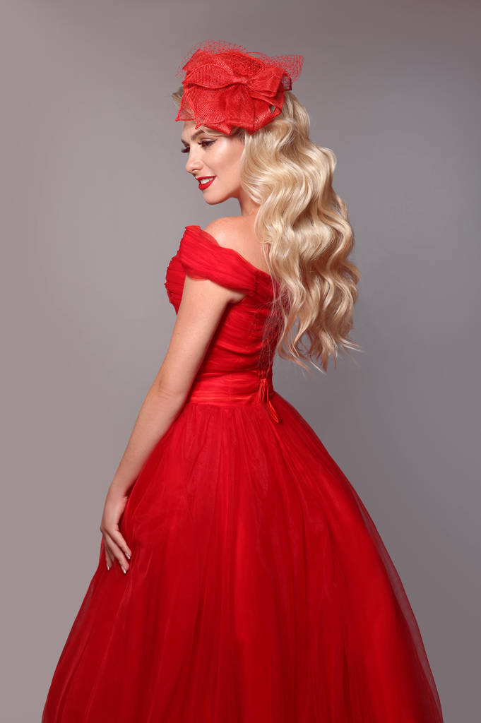 Szőke nő piros ruhában és elegáns kalap elszigetelt stúdió szürke háttér. Gyönyörű szőke lány hosszú hullámos hajjal és sminkkel. Menyasszony stílus. - Fotó, kép