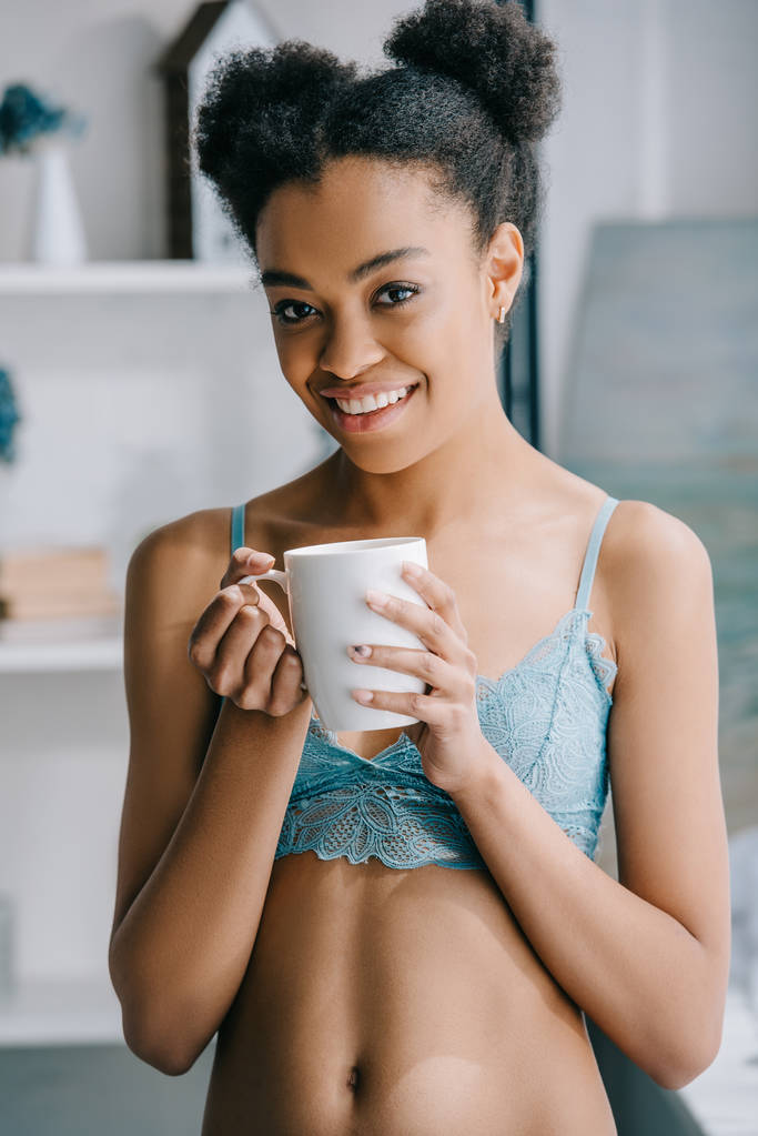 朝の紅茶のカップを保持しているランジェリーで美しい少女は笑みを浮かべてアフリカ系アメリカ人  - 写真・画像