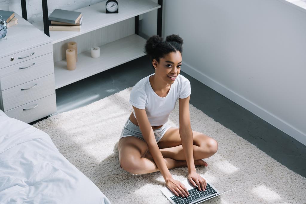 vista ad alto angolo di sorridente ragazza africana americana utilizzando computer portatile bianco seduto al piano in camera da letto
 - Foto, immagini