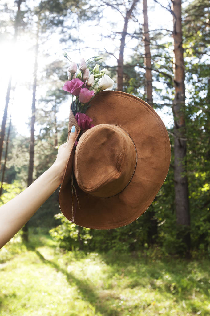 Шляпа и букет розовых цветов в руке женщины на фоне лесной природы
 - Фото, изображение