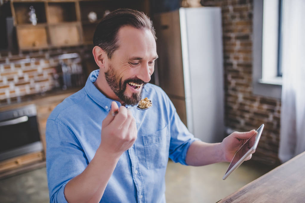 χαμογελώντας γενειοφόρος άνδρας τρώγοντας δημητριακά για το πρωινό και χρησιμοποιώντας ψηφιακό tablet στην κουζίνα - Φωτογραφία, εικόνα