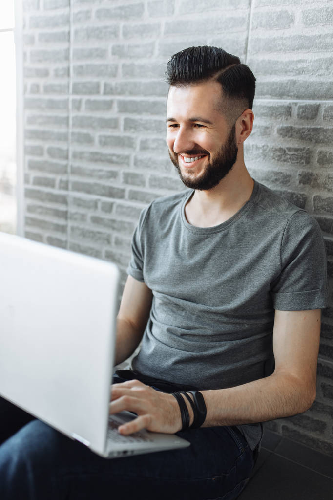 Portrait of a beautiful stylish guy, a hipster, running on a laptop, wearing a gray empty t-shirt, standing on a brick gray wall background. - Photo, Image