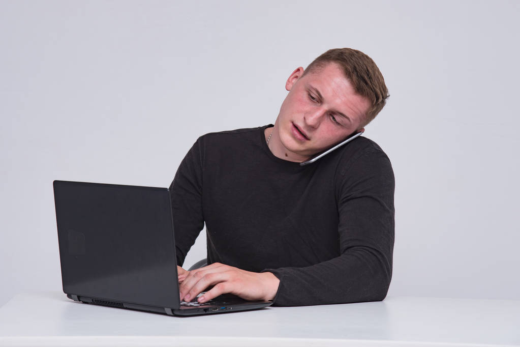 Portrait d'un jeune homme mignon avec un ordinateur portable sur un fond blanc assis à une table avec un smartphone. Il est assis juste en face de la caméra souriant et semble sérieux
 - Photo, image