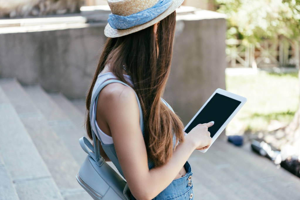 Seitenansicht einer jungen Frau mit Hut mit digitalem Tablet mit leerem Bildschirm auf der Treppe  - Foto, Bild