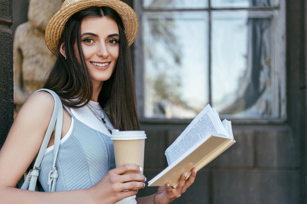 ελκυστική κοπέλα με χαρτί Κύπελλο ανάγνωση του βιβλίου και να χαμογελά στη φωτογραφική μηχανή - Φωτογραφία, εικόνα