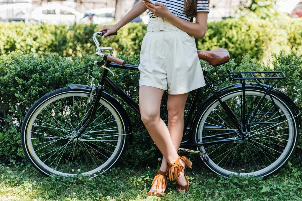 cropped shot of woman with smartphone and retro bicycle standing in park - Photo, Image
