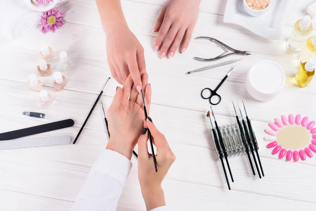 cropped image of beautician doing manicure to woman at table with nail polishes, nail files, nail clippers, cuticle pusher, sea salt, flowers, aroma oil bottles and samples of nail varnishes - Photo, Image