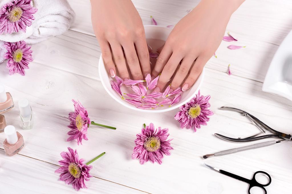 περικοπεί shot γυναίκα λήψη λουτρό με ροδοπέταλα για νύχια στο τραπέζι με λουλούδια, πετσέτες, βερνίκια νυχιών και εργαλεία για μανικιούρ στο σαλόνι ομορφιάς  - Φωτογραφία, εικόνα