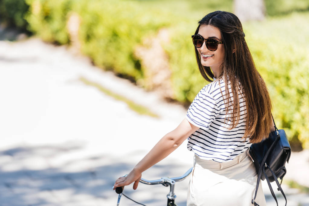 vista lateral de joven mujer sonriente en gafas de sol con bicicleta retro en la calle
 - Foto, Imagen
