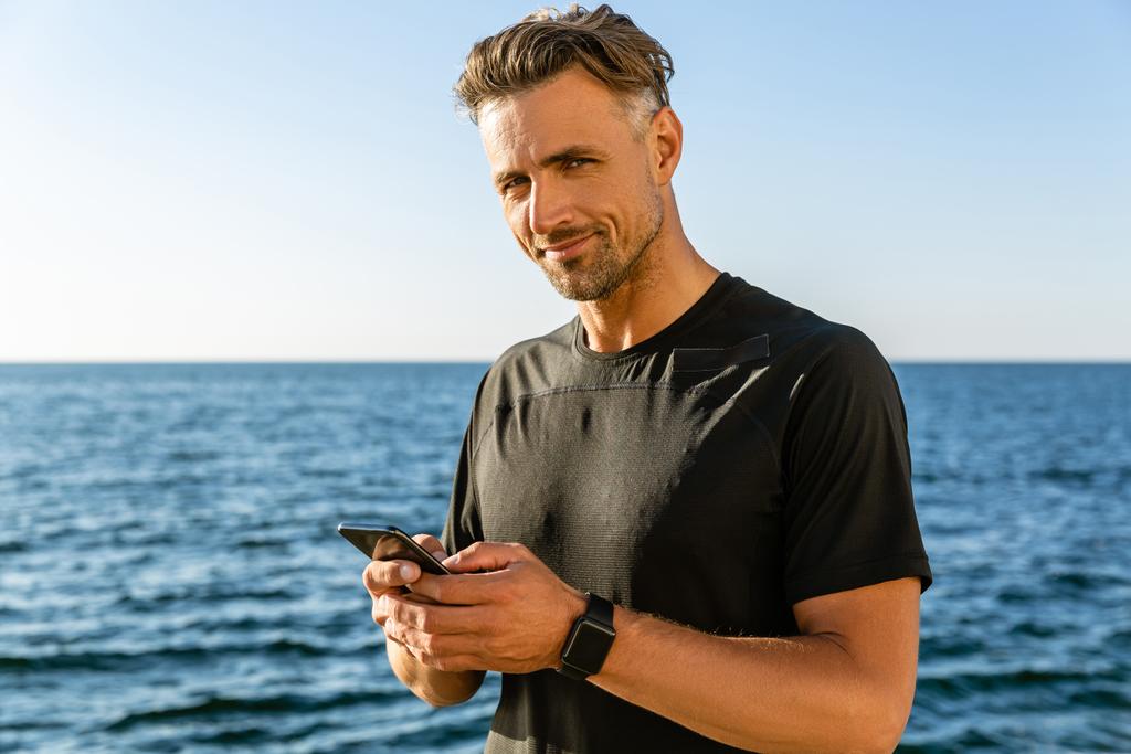 przystojny mężczyzna z smartphone na morza i patrząc na kamery - Zdjęcie, obraz