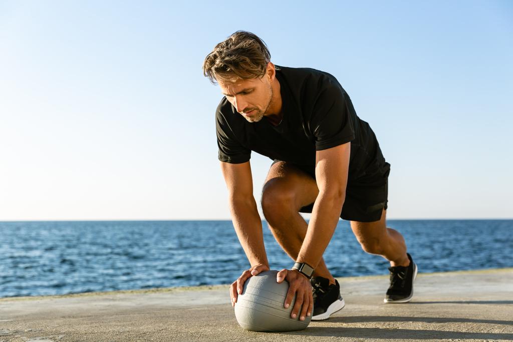 красивый взрослый спортсмен отжимается с мячом на берегу моря
 - Фото, изображение