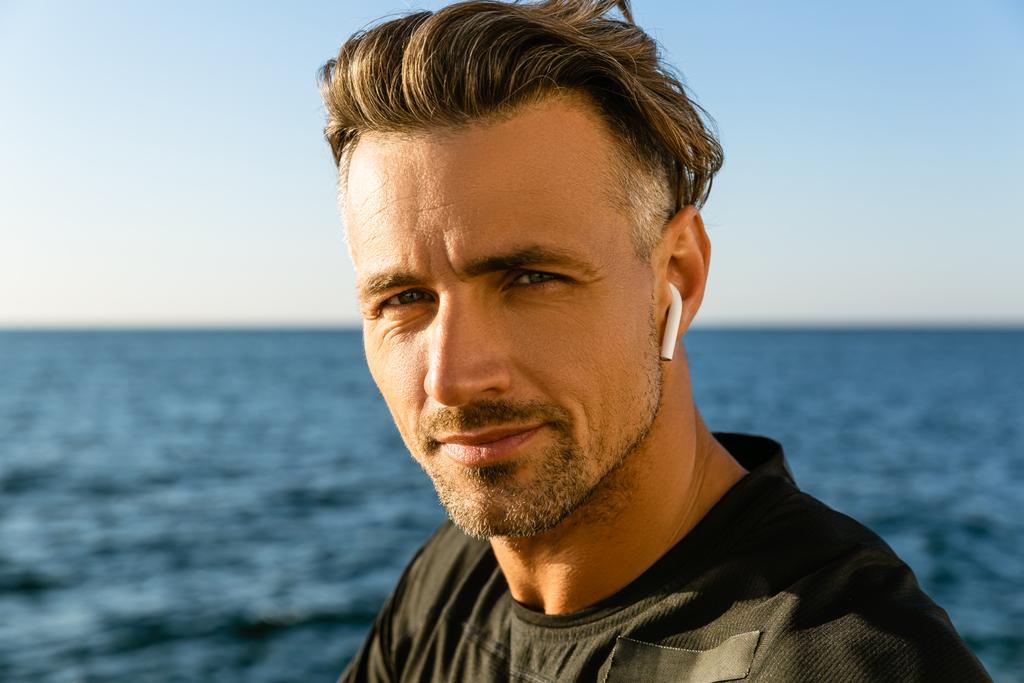 felnőtt férfi tengerparton nézett kamera vezeték nélküli fülhallgató közeli portréja - Fotó, kép