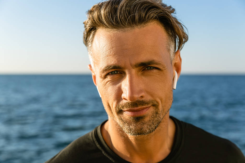 close-up portrait of smiling adult man with wireless earphones on seashore looking at camera - Photo, Image