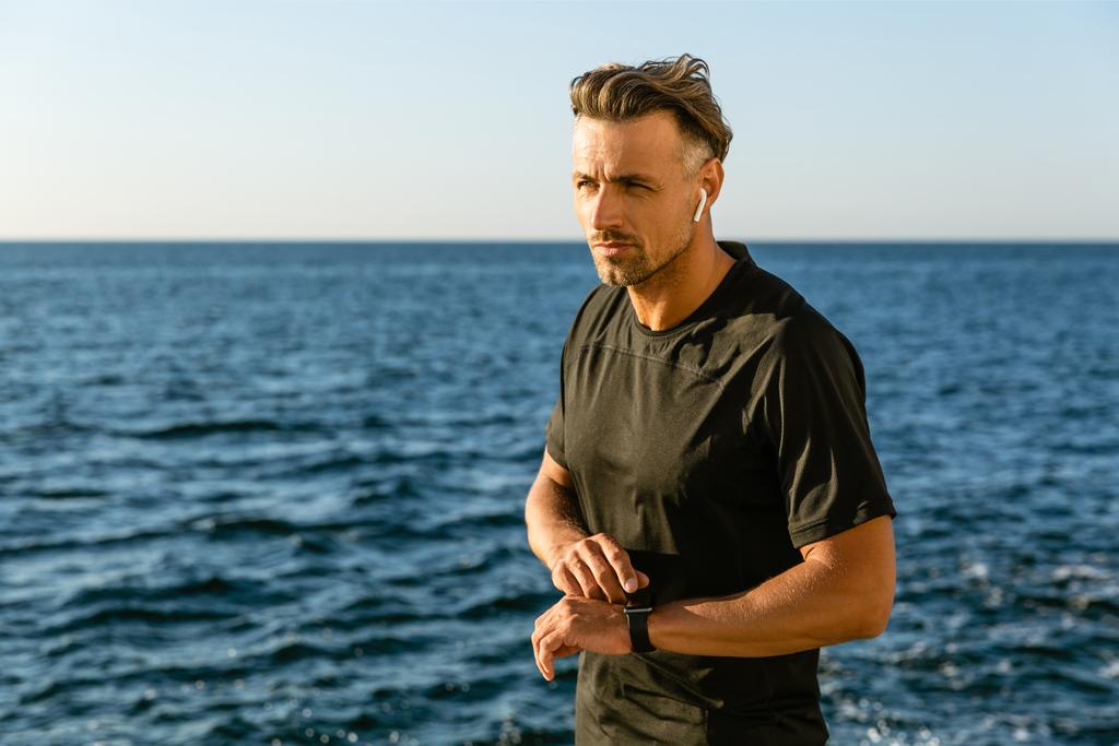 hombre adulto guapo con auriculares inalámbricos y reloj inteligente en la orilla del mar
 - Foto, Imagen