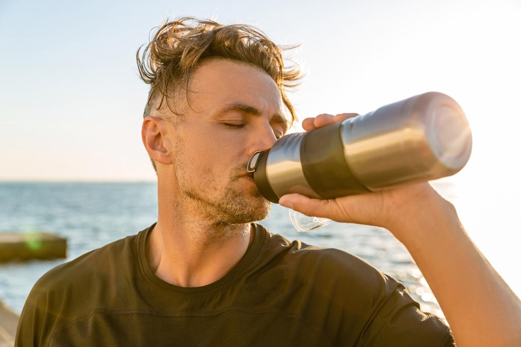 deportista adulto beber agua de la botella de fitness en la orilla del mar en frente de la salida del sol después del entrenamiento
 - Foto, Imagen