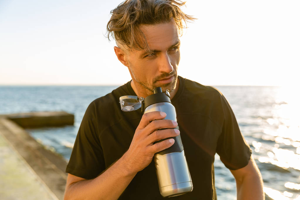 komea aikuinen mies juo vettä fitness-pullosta meren rannalla auringonnousun edessä ja katselee pois
 - Valokuva, kuva