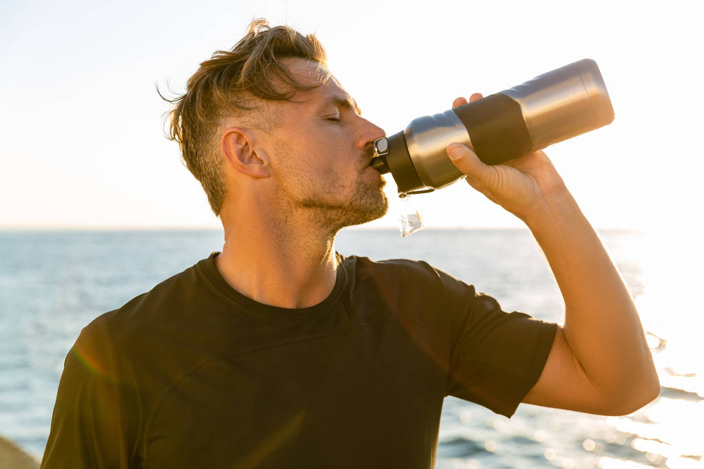 vista lateral del hombre adulto guapo bebiendo agua de la botella de la aptitud en la orilla del mar delante del amanecer
 - Foto, imagen