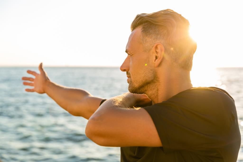 apuesto deportista adulto estirando el brazo antes de entrenar en la orilla del mar frente al amanecer
 - Foto, imagen