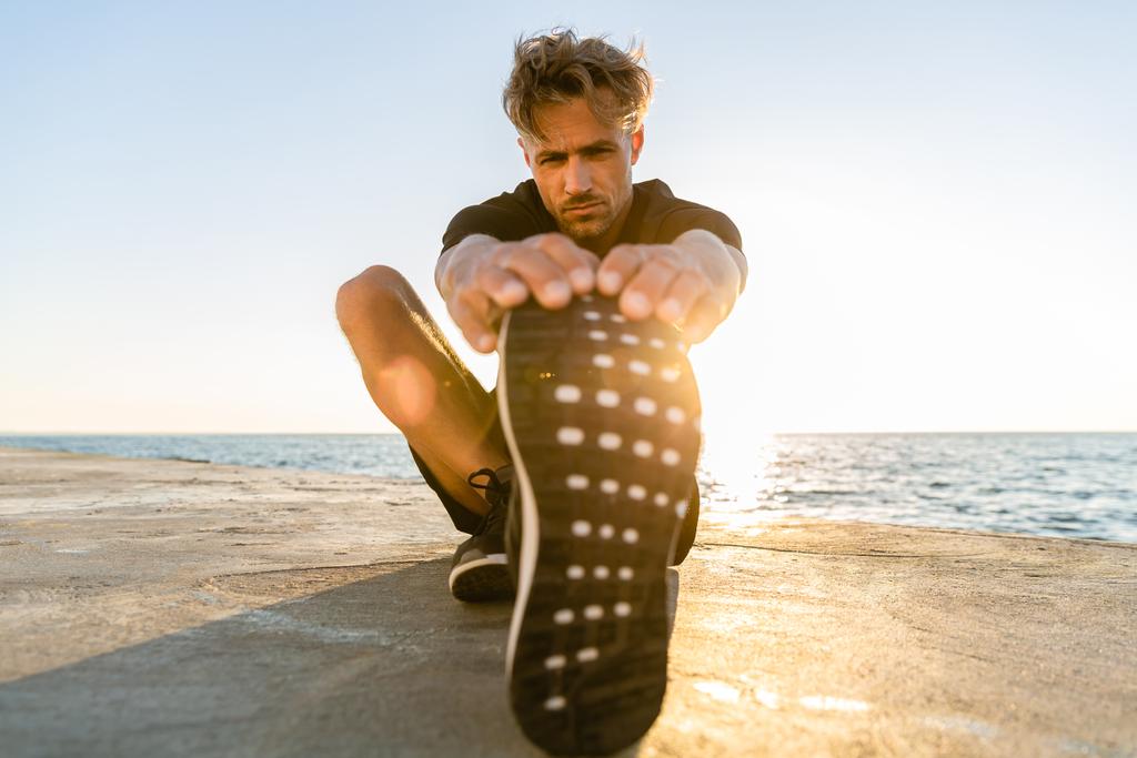 sportowy dorosły człowiek rozciąganie nogi na brzegu morza przed wschodem słońca - Zdjęcie, obraz