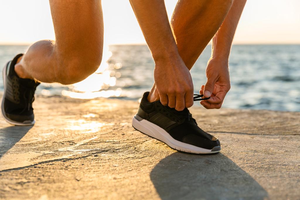 recortado tiro de deportista atando zapatillas de deporte en la orilla del mar delante de la salida del sol
 - Foto, imagen