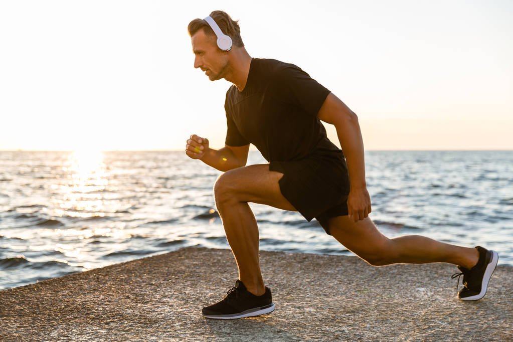 side view of handsome adult sportsman in headphones doing lunges during training on seashore - Photo, Image