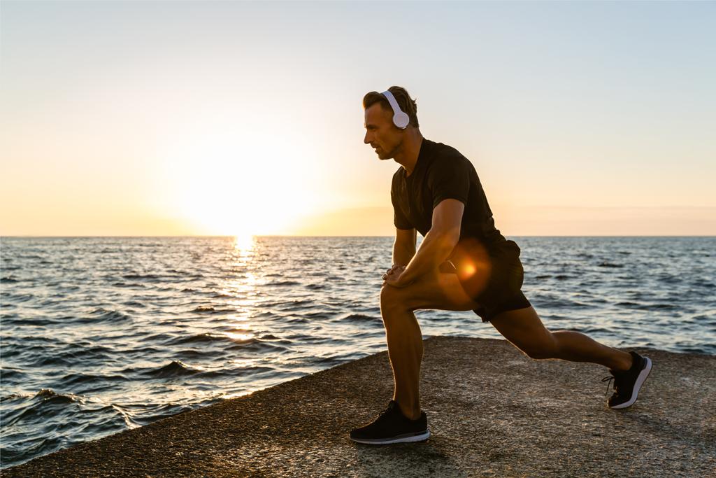 atletische volwassen man in hoofdtelefoons een-legged squats doen tijdens de training aan kust - Foto, afbeelding