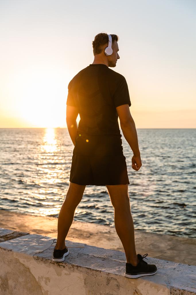 πίσω όψη αθλητική ενήλικα άνδρα ακουστικά που στέκεται στην ακτή μπροστά από την Ανατολή του ηλίου - Φωτογραφία, εικόνα