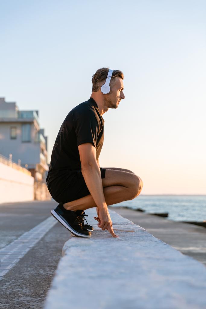 Widok z boku przystojny sportowiec dorosły w słuchawkach stałego przysiady na parapet na seashore - Zdjęcie, obraz