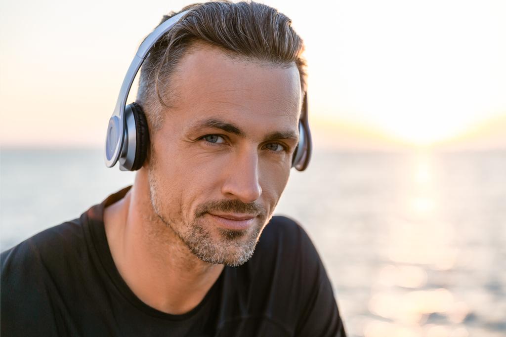 Nahaufnahme Porträt eines attraktiven erwachsenen Mannes mit drahtlosen Kopfhörern, der an der Küste in die Kamera blickt - Foto, Bild