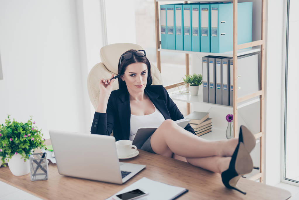 日記と近代的なオフィスのデスクトップに座って戦略の作成の手でペンを保持しているノート パソコンの画面を見ながらテーブルに足を置く思いやりのあるセクシーな女性の肖像画 - 写真・画像