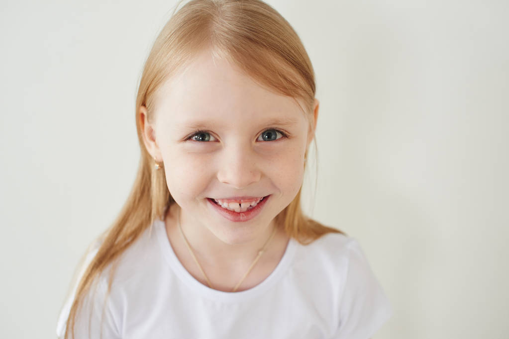 Portrait de petite fille blonde en t-shirt blanc toothy souriant et regardant la caméra sur fond de studio blanc, gros plan
 - Photo, image