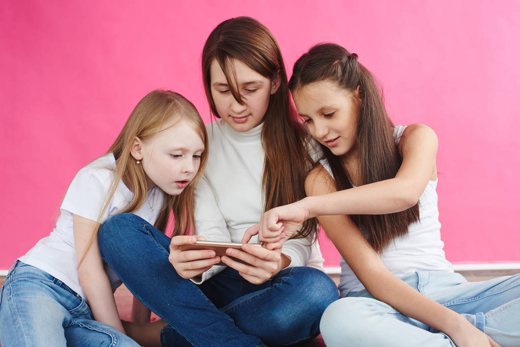 милые девушки в повседневной одежде играют в игры на смартфоне, сидя на розовом фоне студии
 - Фото, изображение