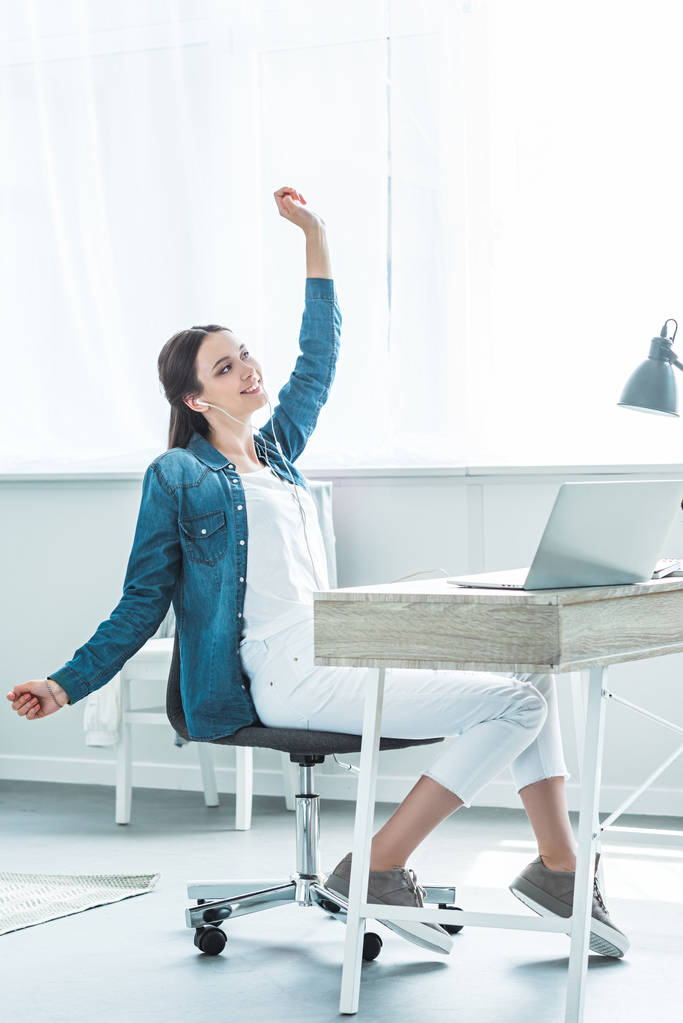 ευτυχισμένος κορίτσι στα ακουστικά αύξηση το χέρι και να ψάχνει μακριά ενώ χρησιμοποιείτε φορητό υπολογιστή στο γραφείο - Φωτογραφία, εικόνα