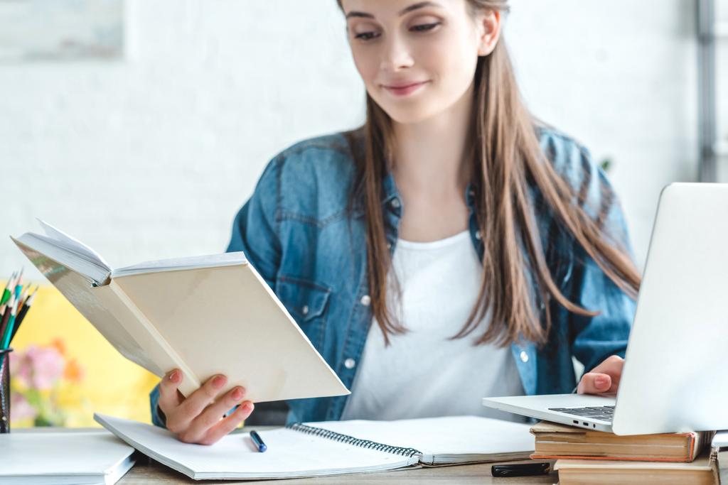 sorridente menina segurando livro e usando laptop enquanto estuda em casa
 - Foto, Imagem