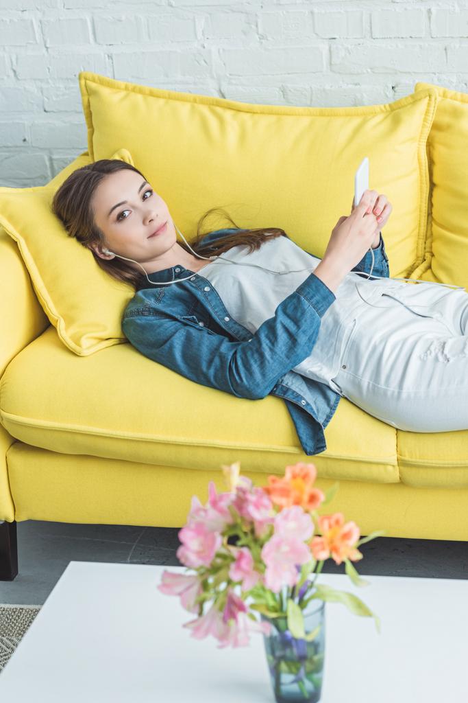 hermosa chica en los auriculares mirando a la cámara mientras está acostado en el sofá y el uso de teléfono inteligente
 - Foto, Imagen