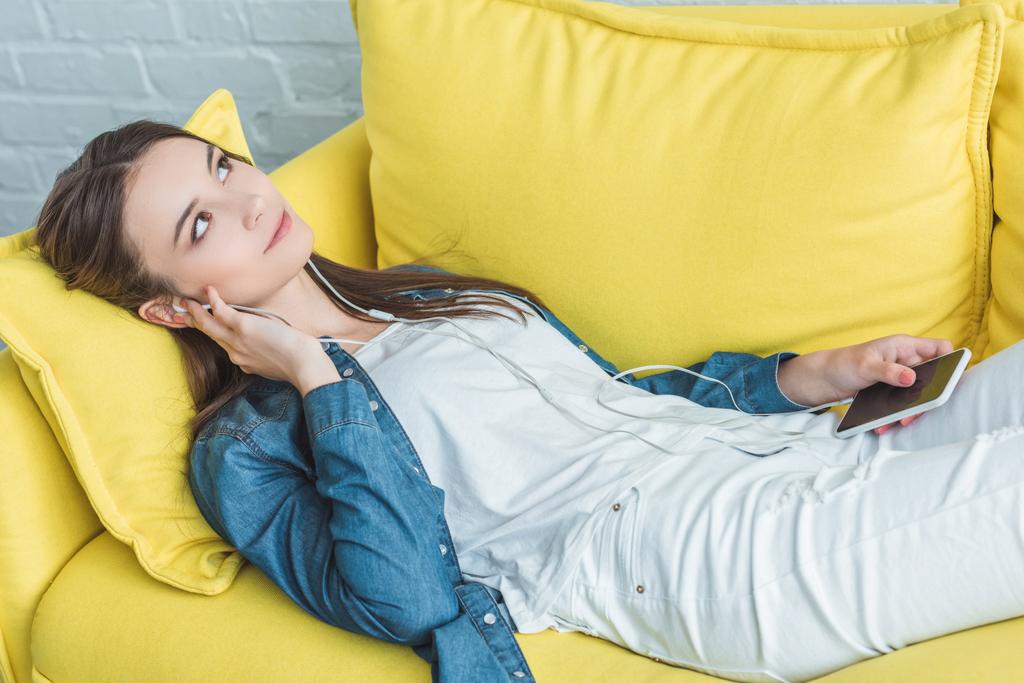 pensive smiling girl holding smartphone and listening music in earphones while lying on sofa and looking up - Photo, Image
