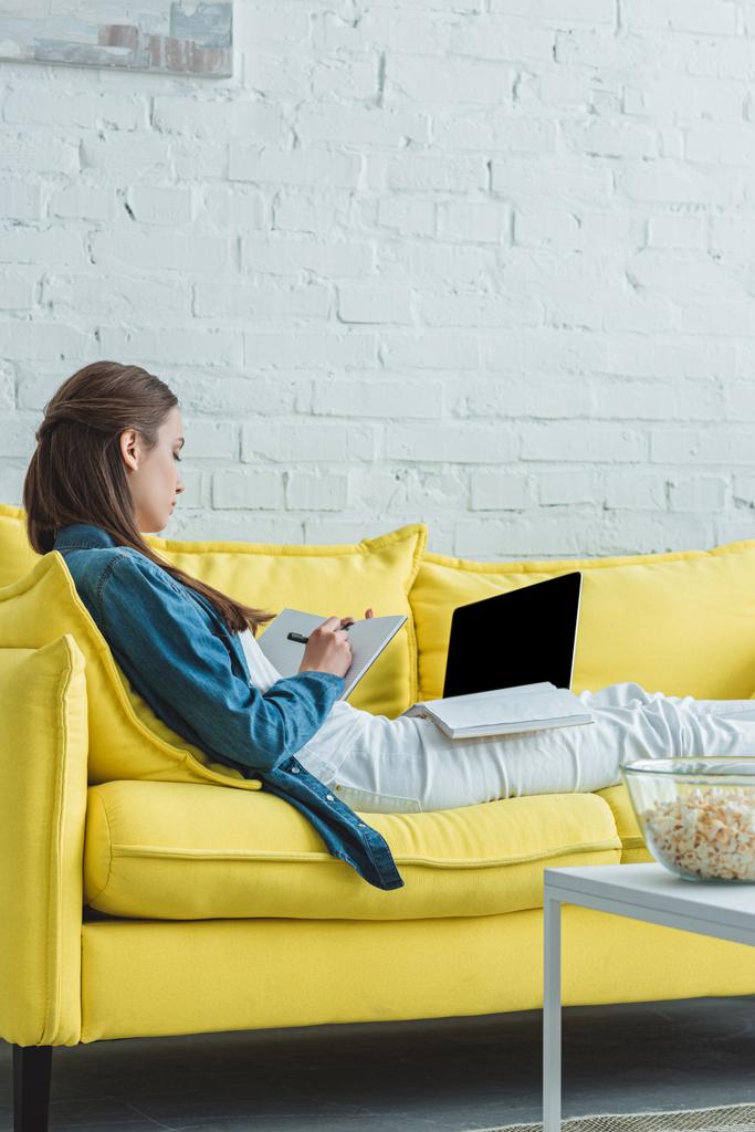 dívka si poznámky do notebooku a pomocí přenosného počítače s prázdnou obrazovkou na gauči doma - Fotografie, Obrázek