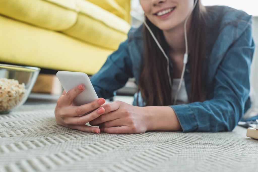 vista parcial de cerca de la chica sonriente en los auriculares usando el teléfono inteligente mientras está acostado en la alfombra
 - Foto, Imagen