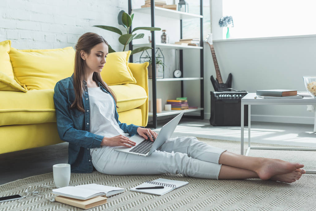 geconcentreerd barefoot meisje met laptop terwijl zittend op een tapijt en thuis studeren - Foto, afbeelding
