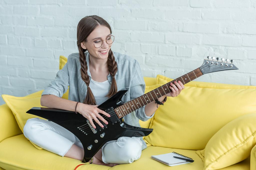 улыбающаяся девочка-подросток, играющая на электрогитаре, сидя на желтом диване с учебником
  - Фото, изображение