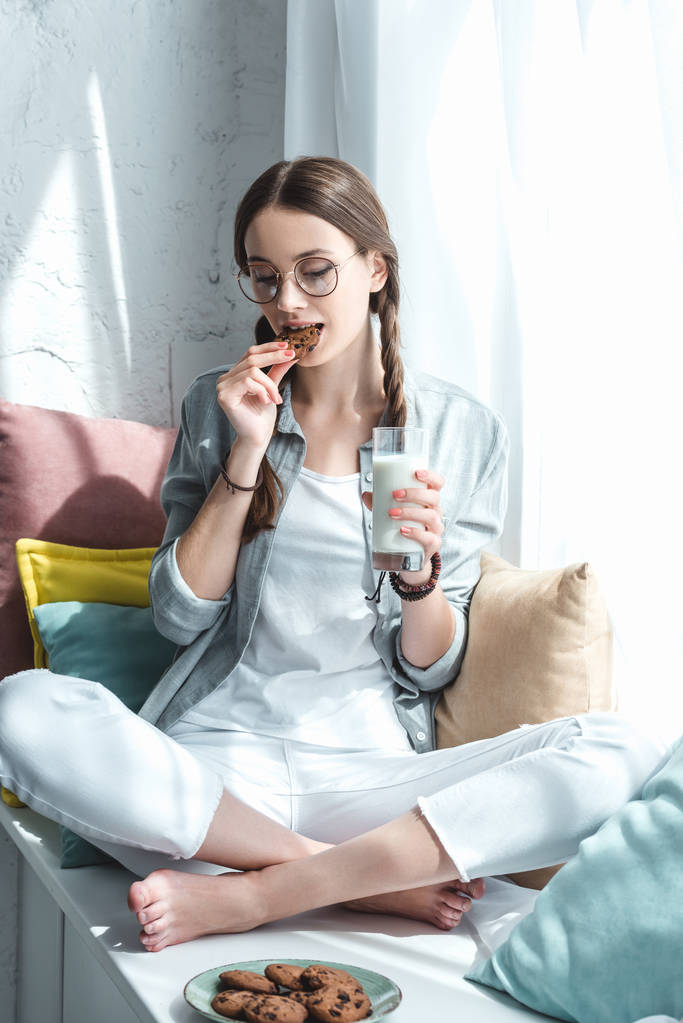 красива дівчина-підліток зі склянкою молока, що їсть печиво на підвіконні
 - Фото, зображення