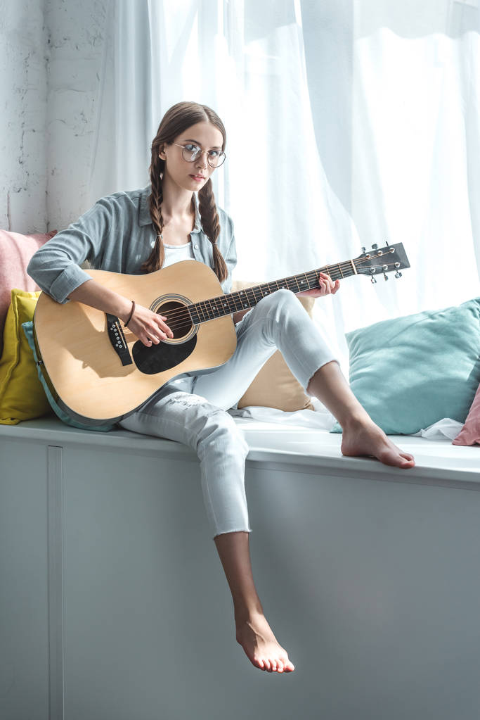 szép tini lány játszik akusztikus gitár, ülve, ablakpárkányon párnával - Fotó, kép